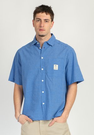 Jeune homme portant une chemise à manches courtes bleu à carreaux avec une poche avant et un pantalon beige, debout devant un fond uni.