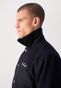 Jūras zila vilnas jaka ar augstu apkakli, ar kontrastējošām baltām pogām un izšūtu detaļu kreisajā krūtī. Gluda tekstūra.