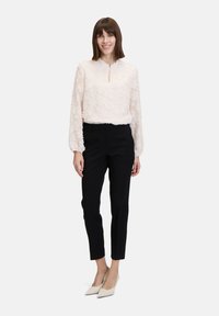 Texturierte, hellrosa Bluse mit einem Schlüssel-ausschnitt und langen Ärmeln, kombiniert mit enganliegenden schwarzen Hosen und beigen spitzen Schuhen.