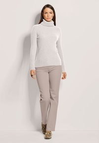 Frau in einem hellgrauen gerippten Rollkragenpullover, beige geraden Hosen und Schlangenleder-Mokassins, die vor einer schlichten Wand steht.