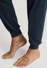 Marine joggers met elastische manchetten, zachte stof, met blote voeten waarop zichtbare tatoeages op de enkels en zolen te zien zijn.