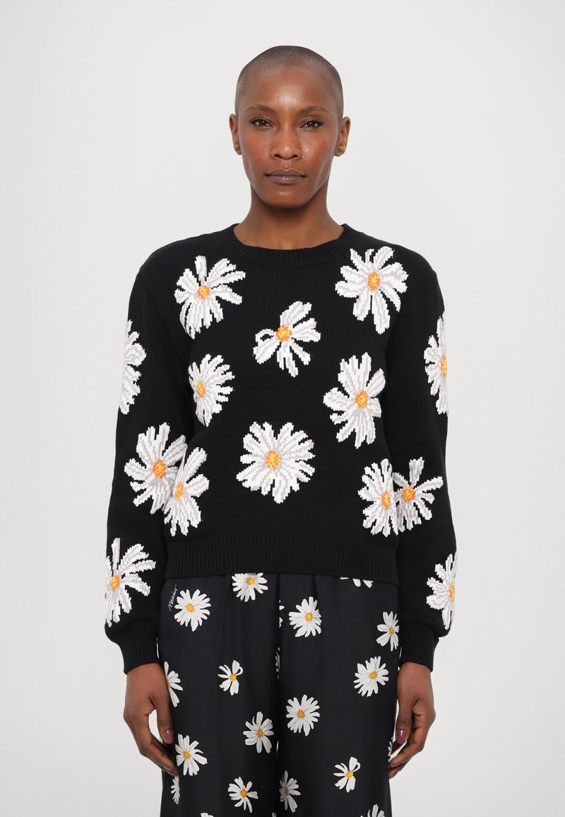 Schwarzer Pullover mit weißer Gänseblümchen-Stickerei und orangefarbenen Mittelpunkten, gerippten Bündchen und Saum. Das Modell trägt passende Hose mit Gänseblümchen-Muster.