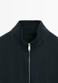 Veste noire avec un col montant et une fermeture éclair argentée partiellement zippée à l'avant, présentée sur un fond blanc.