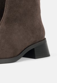 Stivaletto in suede marrone con finitura testurizzata, tacco a blocco e silhouette curvata. Suola in gomma nera con scanalature orizzontali.