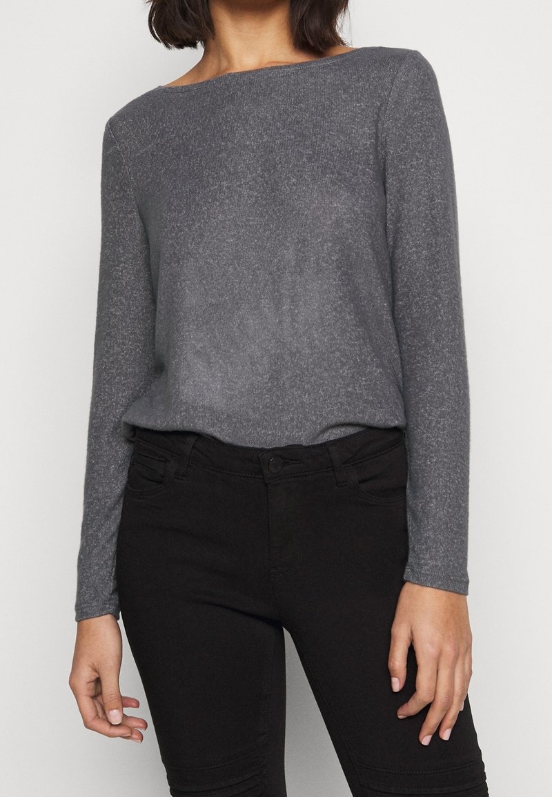 Femme portant un pull gris cintré à manches longues rentré dans un jean noir slim, sur un fond clair uni.