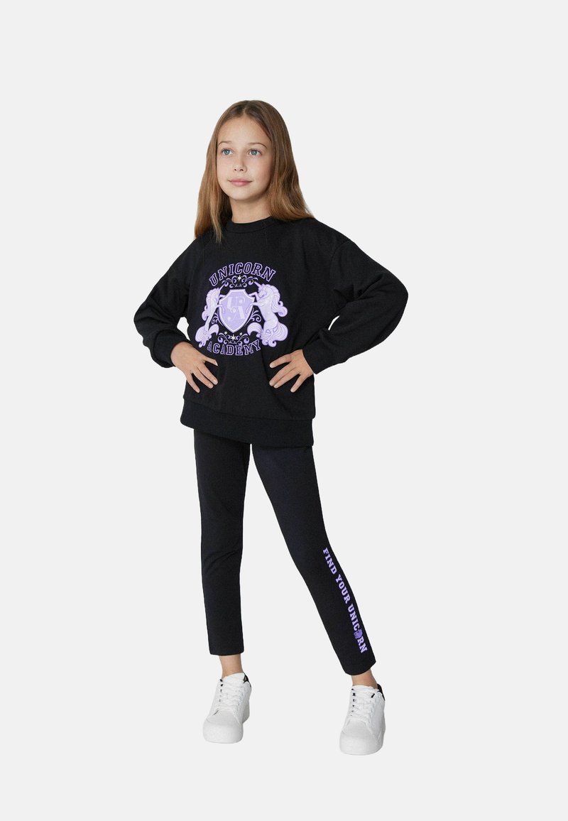 Zwarte sweatshirt met paarse "Unicorn Academy" graphic, gecombineerd met zwarte leggings met de tekst "Find Your Unicorn" en witte sneakers.