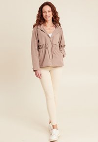 Giacca zip in beige chiaro con vita regolabile, tasche frontali e cappuccio. Indossata sopra un top bianco e abbinata a leggins chiari e sneakers.