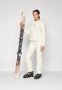 Équipement de ski comprenant un pull en polaire crème, un pantalon de ski beige et des bottes de ski noires. Le personnage tient un ski avec des fixations noires et un design à motifs.