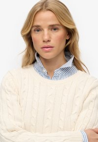 Kremowy sweter w splot kablowy noszony na jasnoniebieskiej i białej koszuli z kołnierzykiem w paski. Posiada okrągły dekolt i ściągacze przy mankietach.