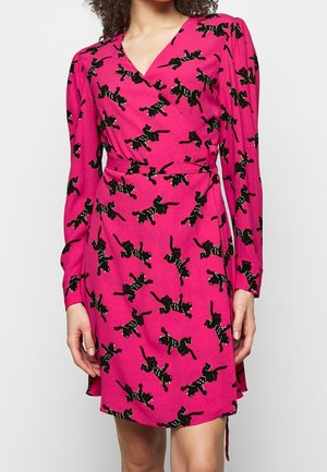 Woman wearing a pink wrap dress with black cat silhouettes, long sleeves, and a waist tie, standing against a plain light background.