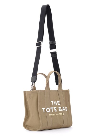 Bolsa de lona beige con correa de hombro negra, que presenta dobles asas superiores y un texto en blanco audaz: "LA BOLSA TOTE MARC JACOBS."