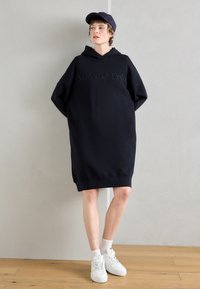 Čierne kapucňové šaty v štýle mikiny s oversized strihom, s vyšitým názvom značky "Marc O'Polo" v tmavšom odtieni na prednej strane.
