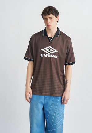 Joven con cabello rizado que lleva una camiseta tipo polo marrón de Umbro y jeans sueltos de color azul claro, sobre un fondo claro y sencillo.