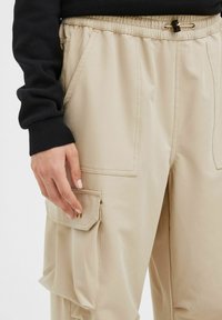 Pantalons cargo beiges avec une taille élastique, poches latérales et une poche à rabat unique. Le tissu semble lisse avec des détails de couture subtils.