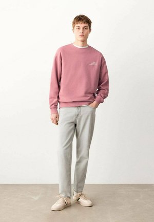 Mladić stoji s jednom rukom u džepu, nosi ružičastu majicu dugih rukava, svijetlosive traperice i bež tenisice na jednobojnoj pozadini.