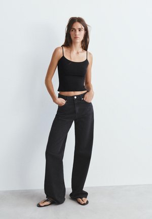 Jonge vrouw staand, draagt een zwart spaghettihemdje, zwarte jeans met hoge taille en wijde pijpen, en zwarte teenslippers op een grijze vloer.