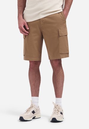 Man wearing beige cargo shorts with side pockets, white socks, and beige sneakers standing against a plain background.