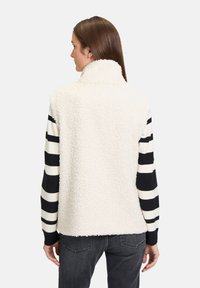 Gilet fluffy color crema con colletto alto; indossato sopra una maglietta a maniche lunghe a righe bianche e nere. Realizzato in tessuto morbido e texture.