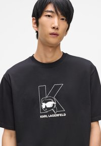 Čierne tričko s krátkym rukávom vyrobené z bavlny, ktoré obsahuje biely grafický dizajn s písmenom "K" a postavou od Karla Lagerfelda.