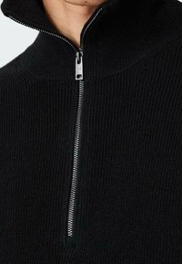 Pull noir en maille avec un col montant et une fermeture éclair centrale argentée. La texture est côtelée, mettant en valeur une coupe ajustée.