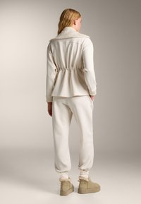 Krémsovo zafarbený outfit s priliehavou sakovkou v peplum štýle s vysokým golierom a elastickým pásom, skombinovaný s voľnými joggermi.
