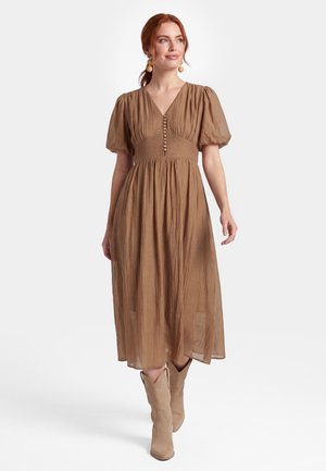 Kvinde med rødt hår iført en brun midi-kjole med pufærmer, knappet foran og beige cowboystøvler, der går fremad.