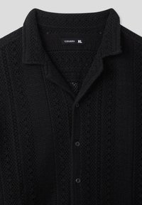 Maglietta nera a maniche corte con un motivo a maglia aperta e testurizzata. Presenta un colletto classico e bottoni frontali. L'etichetta riporta "Terranova XL."