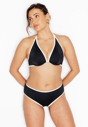 Donna con bikini nero con bordi bianchi, orecchini a cerchio dorati, in piedi con le mani dietro la schiena, guarda di lato contro uno sfondo bianco.