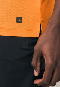 Maglietta sportiva arancione con tessuto strutturato, caratterizzata da un'etichetta con logo nero, abbinata a pantaloncini neri e una mano appoggiata sul tessuto.