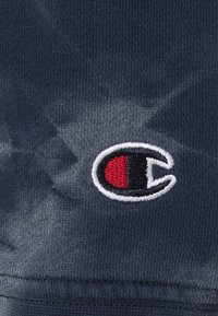 Tmavě modrá bavlněná látka s batikovým vzorem; vyšitá logo s červenými, černými a bílými akcenty poblíž spodní části.