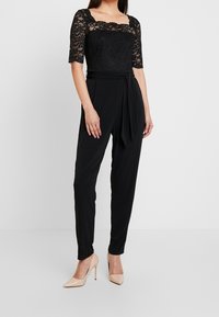 Nainen tiukassa mustassa haalarissa, jossa on pitsihihat, seisoo neutraalin taustan edessä, jalassa nude-korkokengät.