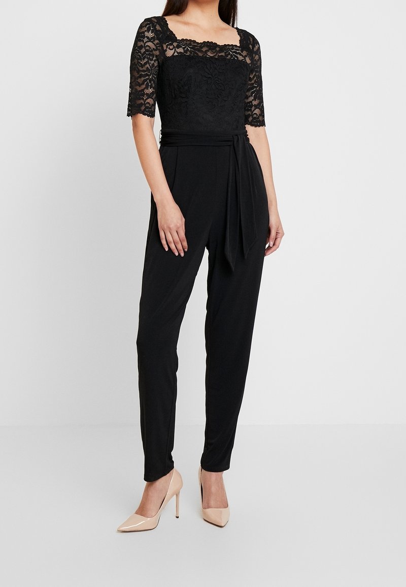 Nainen tiukassa mustassa haalarissa, jossa on pitsihihat, seisoo neutraalin taustan edessä, jalassa nude-korkokengät.