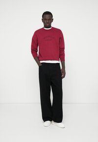 Sweatshirt bordeaux avec "Originals" imprimé en noir, assorti à un pantalon large noir et des baskets blanches. Fond uni.