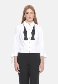 Camicia bianca con design di cravatta a farfalla nera, bottoni dorati e maniche lunghe con polsini spezzati. Indossata con pantaloni neri.