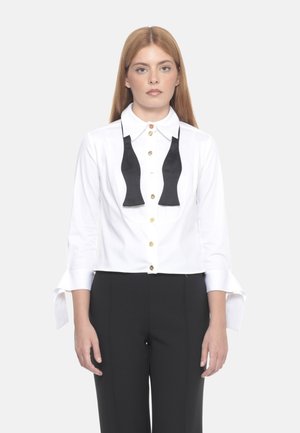Camicia bianca con design di cravatta a farfalla nera, bottoni dorati e maniche lunghe con polsini spezzati. Indossata con pantaloni neri.