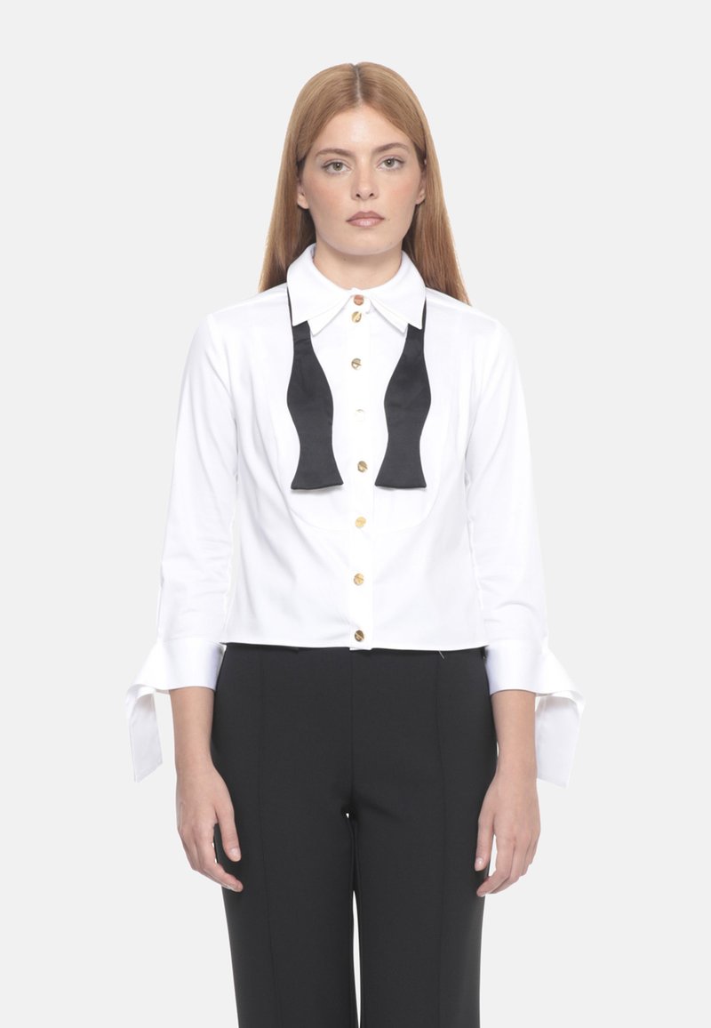 Camicia bianca con design di cravatta a farfalla nera, bottoni dorati e maniche lunghe con polsini spezzati. Indossata con pantaloni neri.