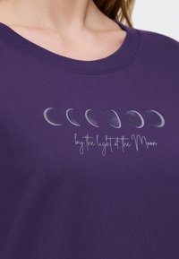 Fioletowa bawełniana koszulka z okrągłym dekoltem, z grafiką faz Księżyca nad napisem "przy świetle Księżyca" w białym kursywie.