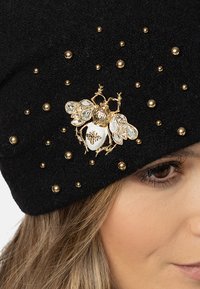 Căciulă din fetru negru, decorată cu o aplicație de albină din aur și cristale clare, alternând cu încuietori de aur, cu un design texturat și o potrivire strânsă în jurul capului.