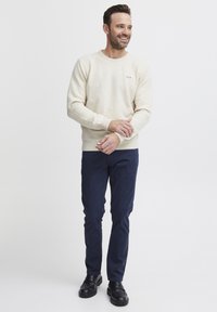 Beige kötött pulóver kerek nyakkal és kis logóval. Sötétkék, slim-fit nadrág és fekete cipő. Egyszerű, letisztult vonalak és textúrák.
