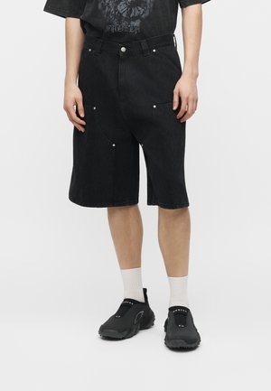 Άτομο που φοράει μαύρα τζιν σορτς μέχρι το γόνατο, μαύρα slip-on αθλητικά παπούτσια, λευκές κάλτσες και μαύρο γραφικό T-shirt, στέκεται μπροστά σε λευκό φόντο.