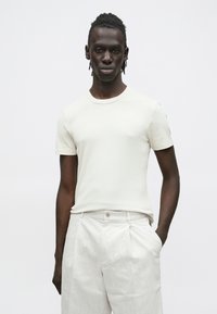 Jovem homem negro com cabelo trançado veste uma camisa canelada de manga curta em tom off-white e calças plissadas bege claro, posando contra um fundo branco.