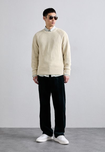 Suéter de canalé color beige claro, camisa con cuello azul claro debajo, pantalones de pierna ancha negros, zapatillas blancas y gafas de sol tipo aviador.