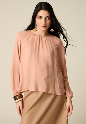 Mujer con cabello largo y oscuro, vistiendo una blusa suelta color durazno, falda beige, pendientes grandes con motivos florales y pulseras gruesas en una muñeca.