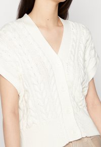 Vit stickad kofta med V-ringning, korta ärmar och ett texturerat kabelmönster med fyra knappar fram.