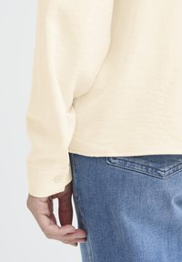 Light beige long-sleeve shirt with a soft texture, featuring a buttoned cuff. Worn with blue denim jeans showing a back pocket detail.