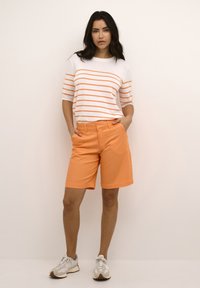 Camisa blanca con rayas naranjas, combinada con pantalones cortos naranjas con bolsillos y un diseño de corte recto. Se lleva con zapatillas ligeras.