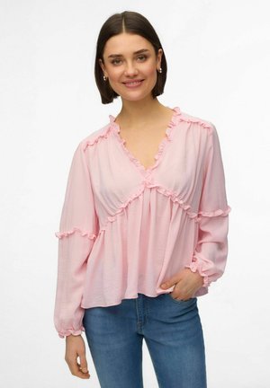 Femme aux cheveux bruns courts portant un chemisier rose clair à volants et un jean bleu, souriant avec une main dans la poche, sur fond blanc.