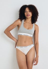 Sivý bavlnený športový podprsenka s čiernym pásom "Calvin Klein". Biela bavlnená tangá s podobným pásom. Modelka pózuje s jednou rukou na páse.