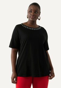Camiseta negra de manga corta con escote redondeado que presenta tachuelas plateadas decorativas alrededor del cuello. Tejido suave, corte relajado. Pantalones rojos.