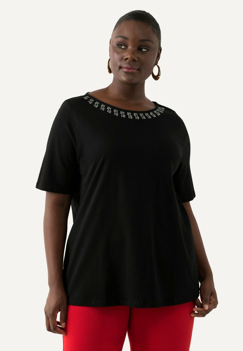 Camiseta negra de manga corta con escote redondeado que presenta tachuelas plateadas decorativas alrededor del cuello. Tejido suave, corte relajado. Pantalones rojos.